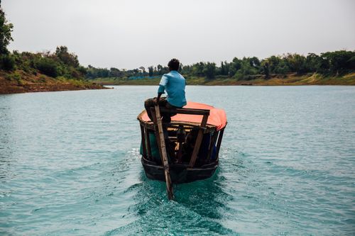 Free A man steers a wooden boat on a serene river surrounded by lush greenery on a sunny day. Stock Photo