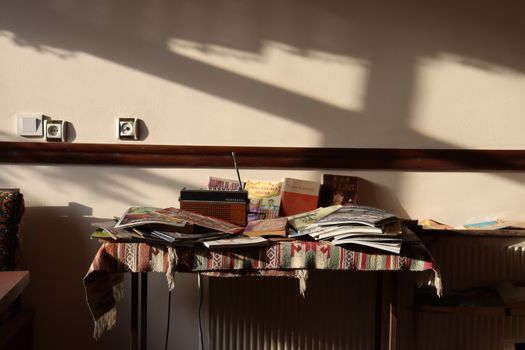 A collection of vintage books and a radio on a sunlit table with a woven cloth.