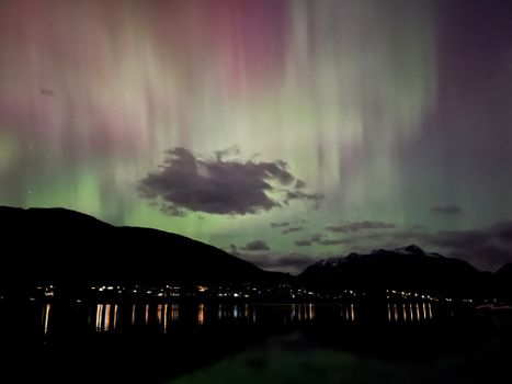 Vibrant aurora borealis illuminating the night sky above a tranquil lake and mountain silhouette.