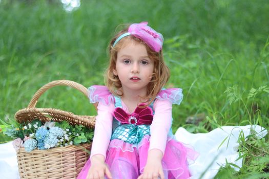 A young girl dressed as a mermaid sits on grass with a wicker basket of flowers. Captured in daylight.