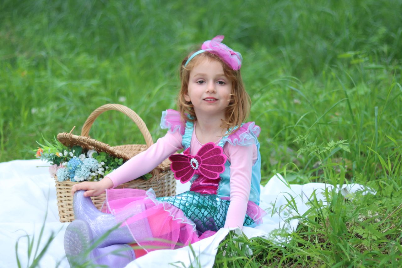 young girl dressed as a mermaid sits in a grassy field with a picnic b