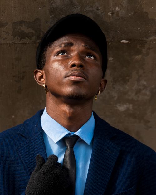 Free Portrait of a young man in a blue suit and hat looking upward against a textured wall. Stock Photo