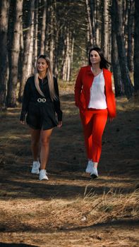 Two women in stylish outfits walking on a sunny forest path, showcasing fashion in nature.