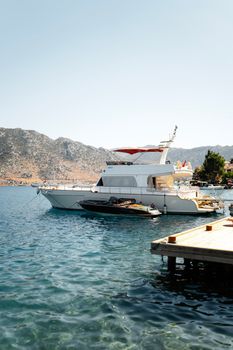 A luxury yacht docked at a serene harbor with clear blue water and scenic mountain backdrop.