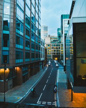 View of a modern London street with buildings at dusk, featuring a cyclist and cars.