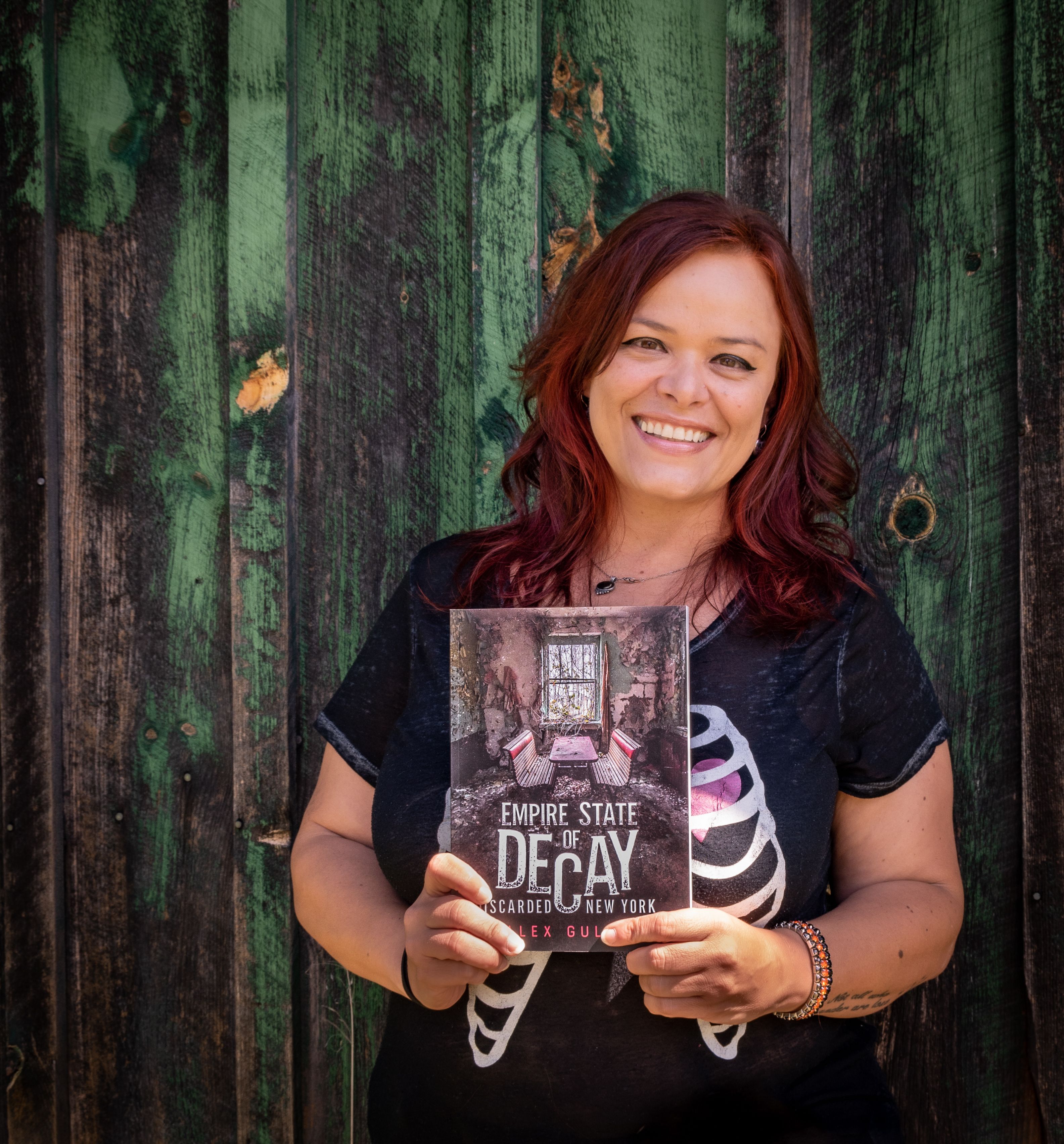 Woman smiling and holding 'Empire State of Decay' book against a wooden background outdoors.