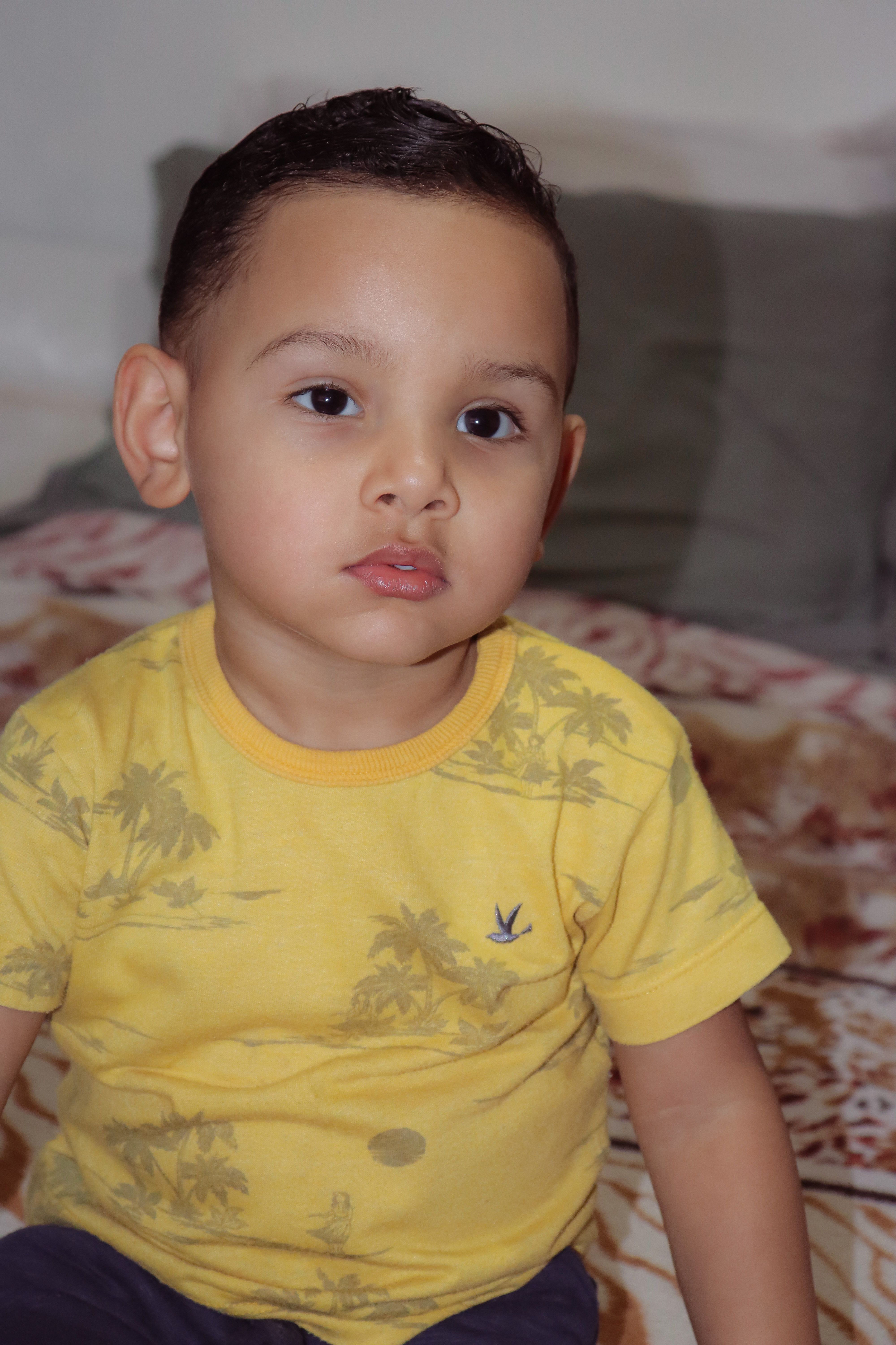 Charming toddler sitting indoors in a yellow t-shirt with a pensive expression.