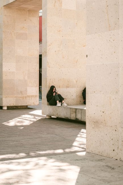 A person sits alone on a marble bench in an urban architectural setting, bathed in sunlight.
