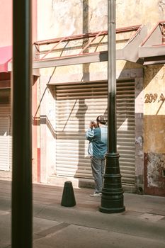 Man taking photographs on a sunny urban street near a shuttered storefront.