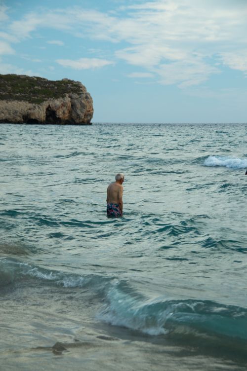 Free A lone swimmer wades through peaceful ocean waters near a rocky coastline under a partly cloudy sky. Stock Photo