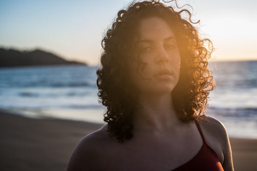 A woman with curly hair gazes thoughtfully on a beach during sunset, capturing a serene moment.