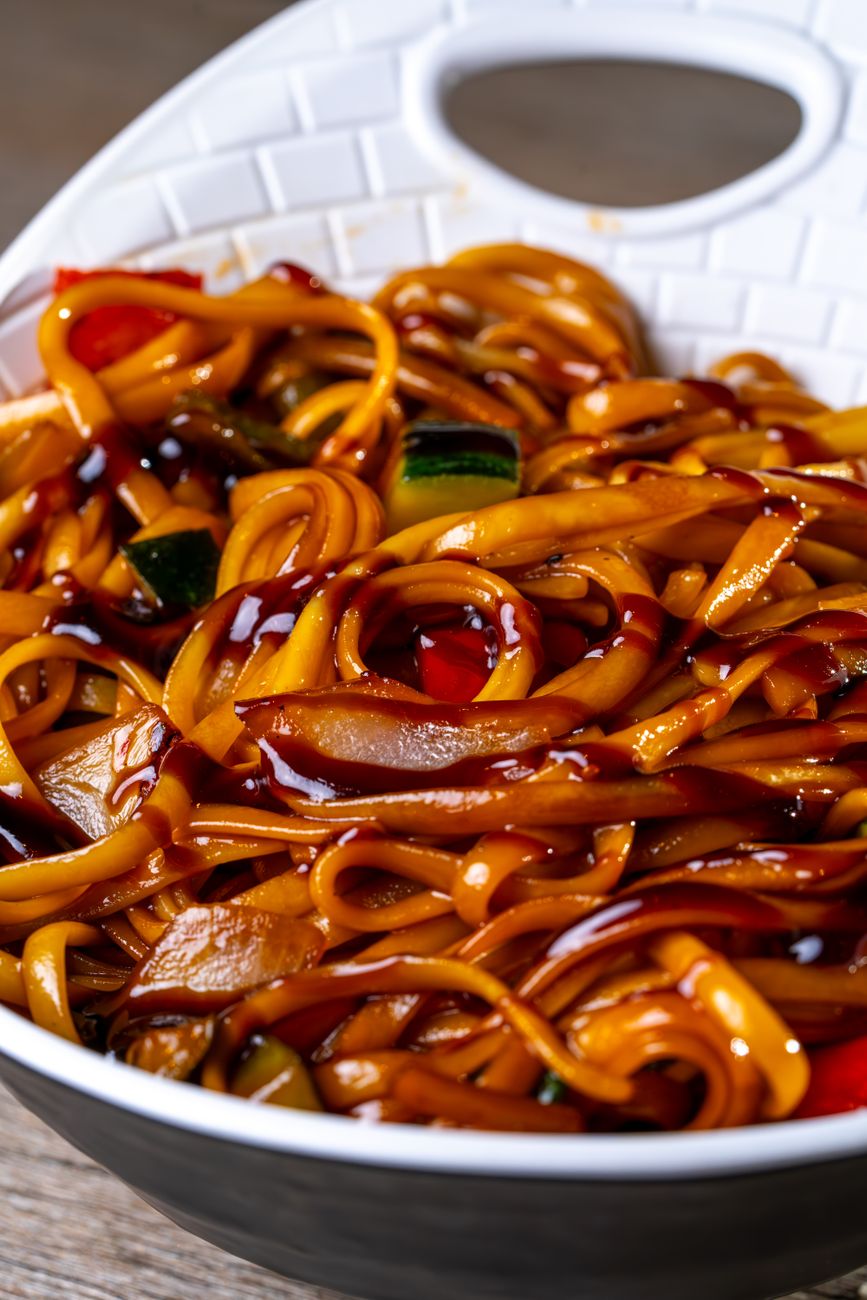 Dark, glossy hakka noodles tossed with colorful vegetables in a wok, the noodles glistening with spicy chili-infused sauce and steam rising from the intense heat