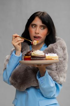 A woman in a fur wrap and blue suit indulges in assorted cupcakes, savoring the flavor.