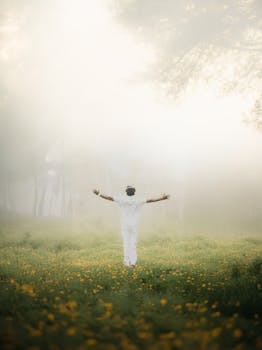 A person standing with open arms in a misty meadow filled with yellow flowers.