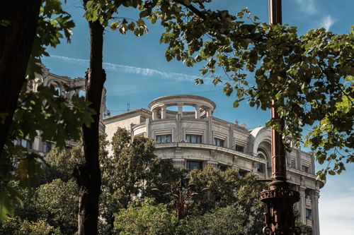 Free A scenic view of a historic building in Bucharest framed by lush green trees. Stock Photo