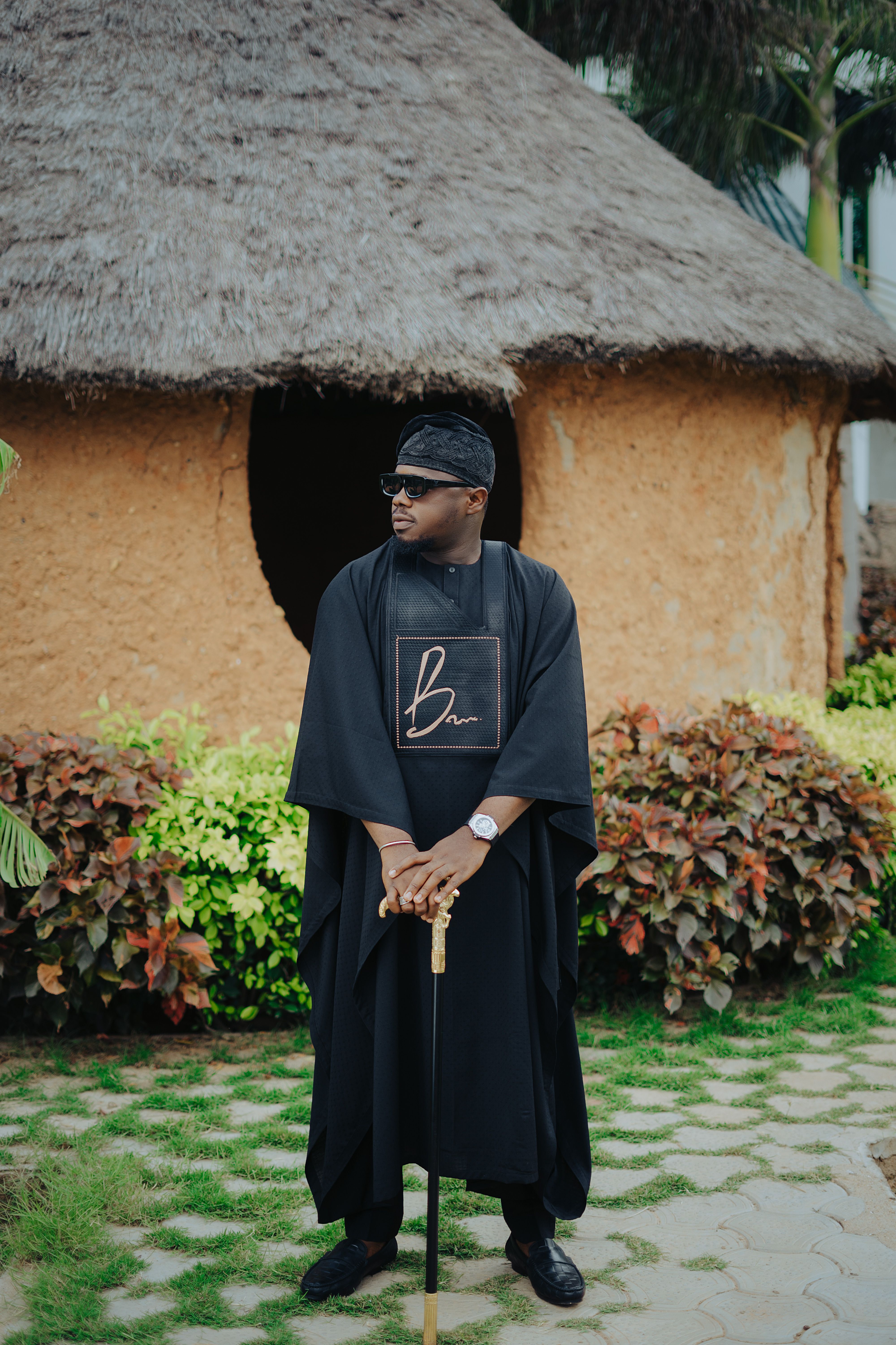 Man standing in garden wearing traditional attire, holding a cane.