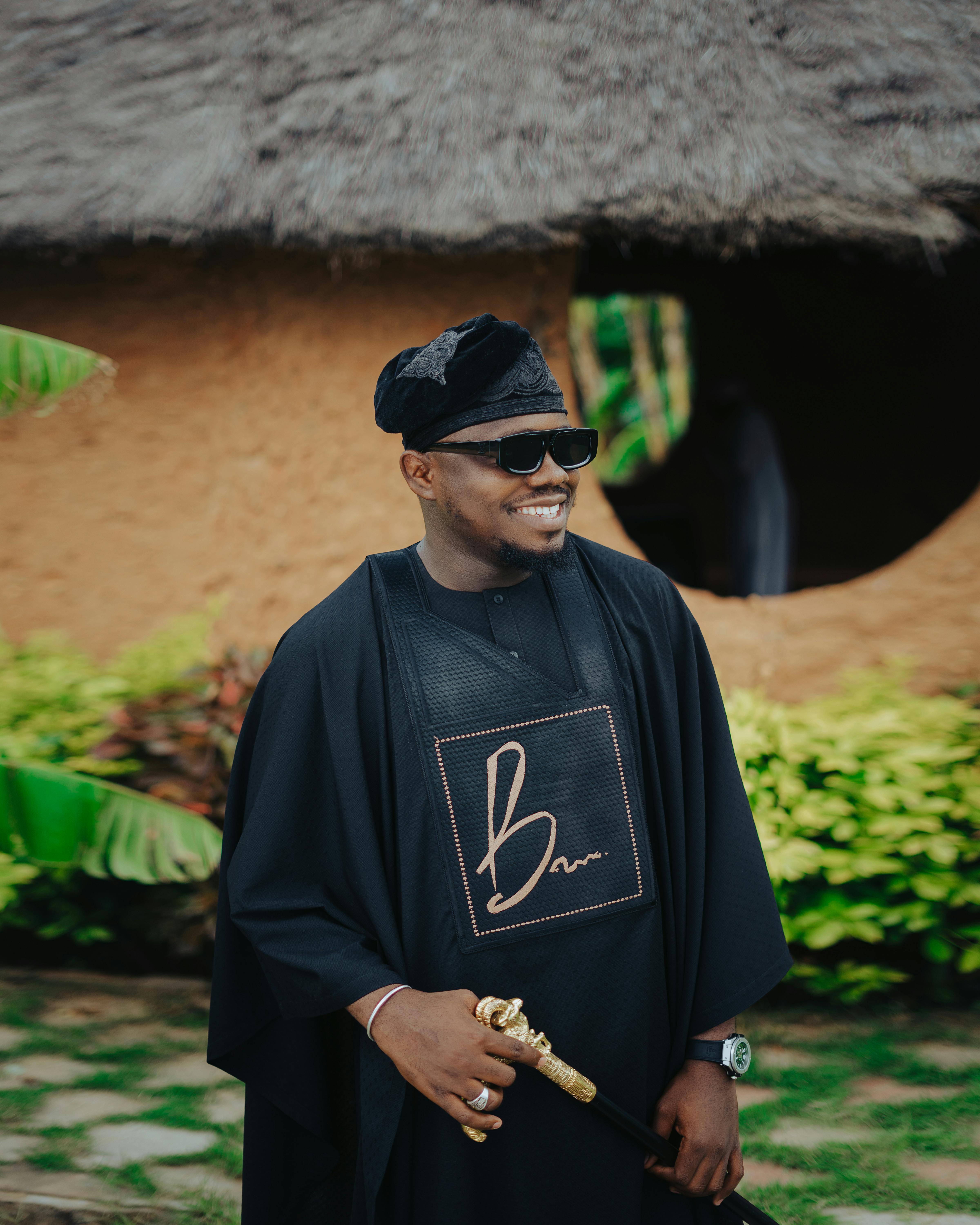 Man wearing traditional black attire with hat and sunglasses, smiling outdoors.