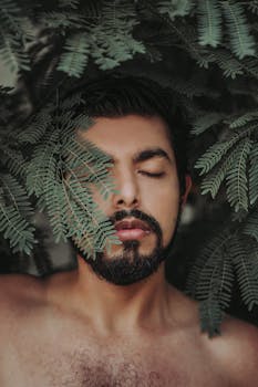 这是一幅特写肖像，画中男子闭着双眼，周围环绕着郁郁葱葱的绿叶