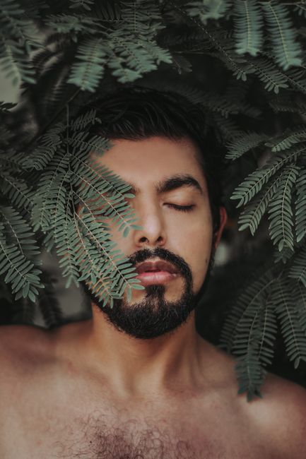 Close-up portrait of a man with closed eyes surrounded by lush green leaves.