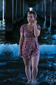 Woman poses under a pier in a floral dress with water and waves around her, capturing a serene summer scene.