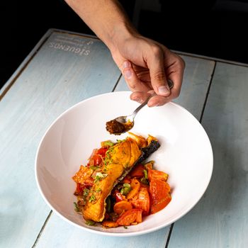 Hand adding spices to gourmet salmon dish with fresh vegetables on rustic table.