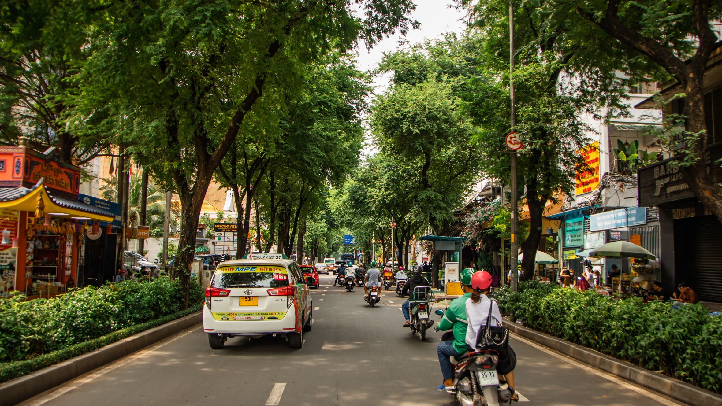 Vibrant street view in Ho Chi Minh City with taxis and motorbikes under lush trees.