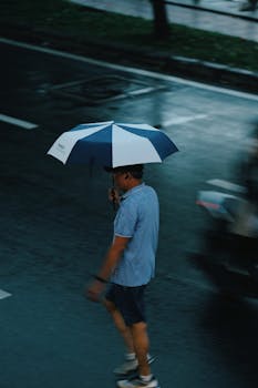 A man walks in the rain holding a blue and white umbrella.
