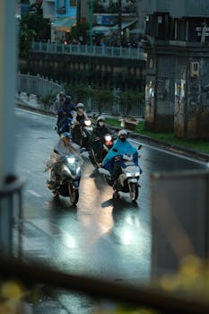 Motorcyclists navigate a wet urban road during rain, wearing helmets and rain gear.