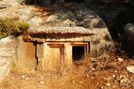 An ancient rock-cut tomb entrance hidden in a rocky, arid landscape under bright sunlight.