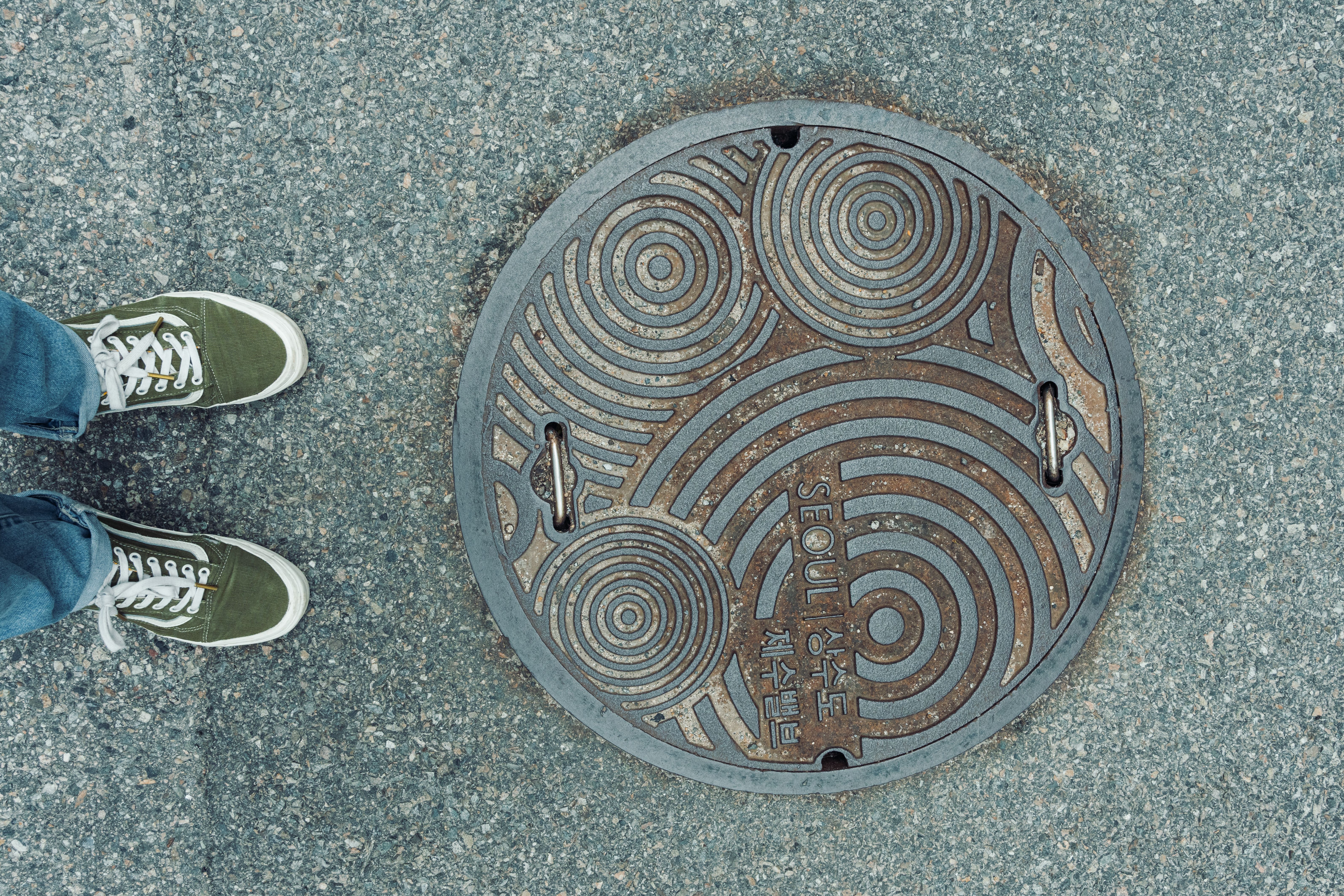 Artistic sewer cover design on a city street, with a person's green sneakers stepping on it.