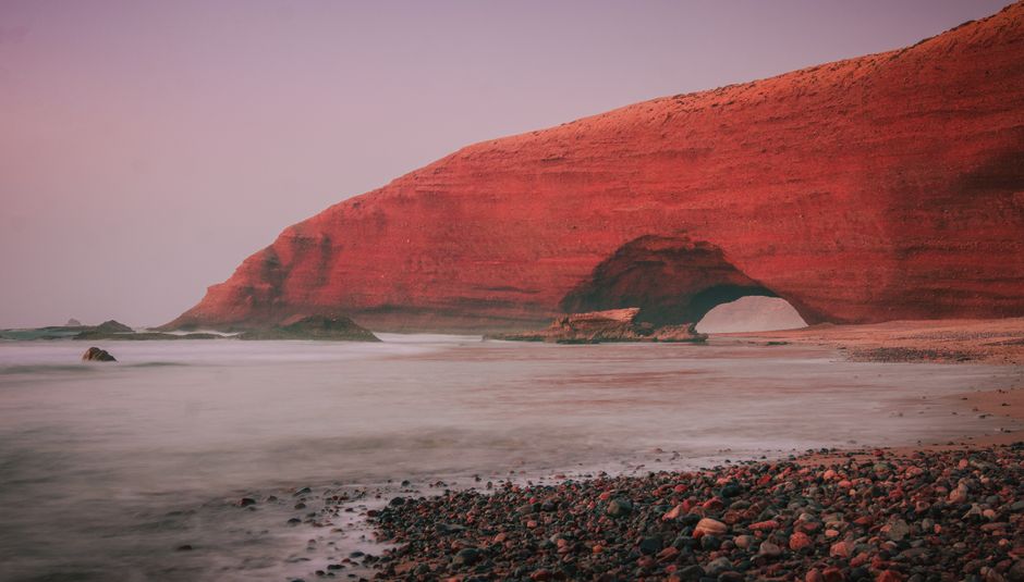 Stunning view of a large red rock arch on a misty beach during twilight, creating a serene atmosphere.