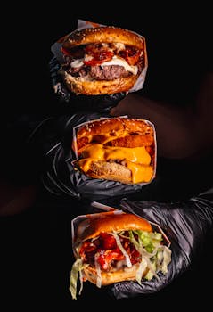 Three gourmet cheeseburgers with various toppings held in black gloved hands on a dark background.