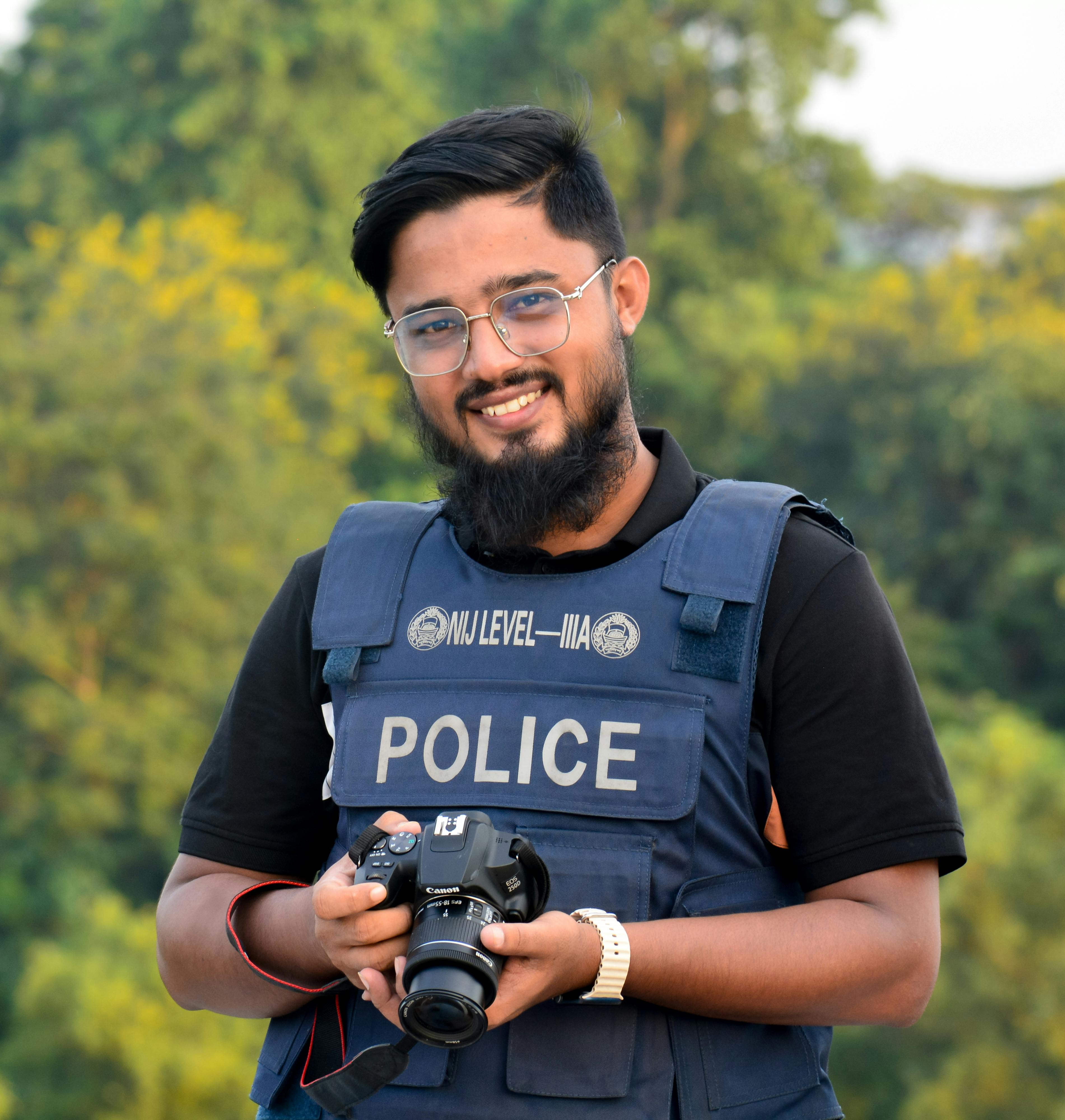 Bangladeshi police officer smiling and holding a camera outdoors. Captured in natural light.