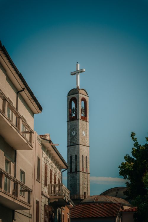 Free A tall church bell tower with a cross stands against a clear blue sky, surrounded by buildings. Stock Photo