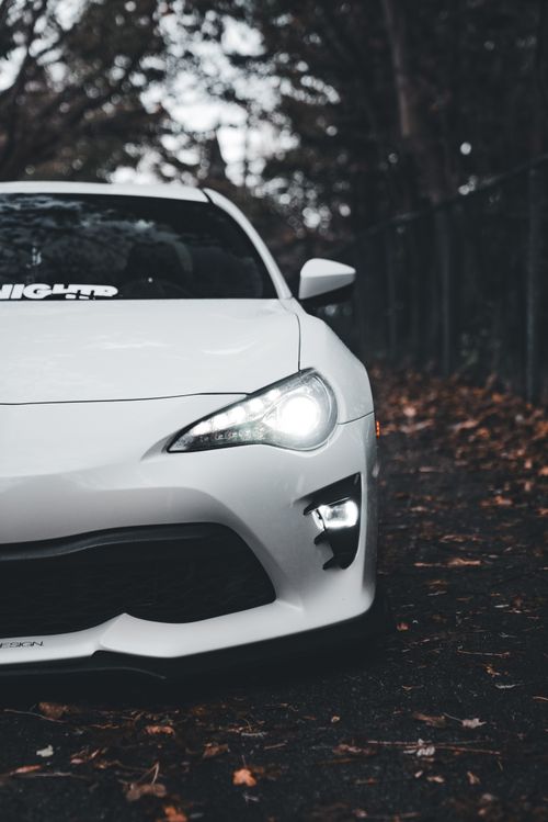 Free White sports car with headlights on, parked on a leaf-strewn road during autumn Stock Photo