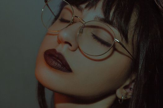 Artistic portrait of a woman with glasses and dark lipstick in low light.