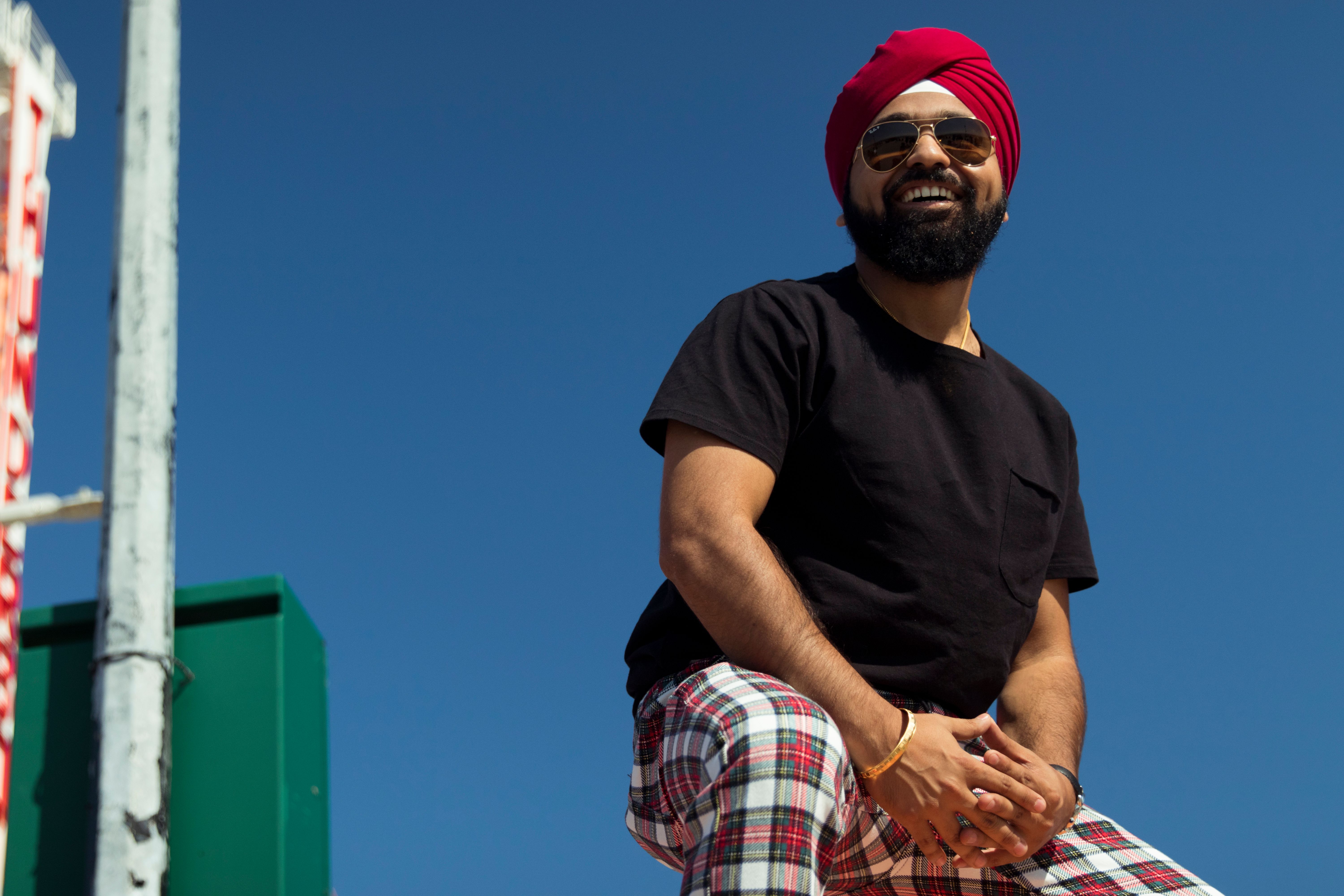 A man in a red turban and sunglasses poses under a clear blue sky, smiling.