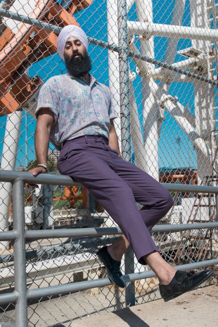 Man in casual summer attire sitting by a wire fence in an urban outdoor area.