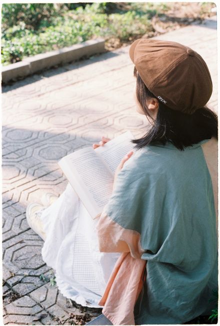 A young woman enjoys reading outdoors, sitting on a sunny sidewalk.