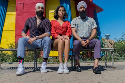 Free Three stylish adults seated outdoors in front of a colorful structure. Sunny day with bright outfits. Stock Photo