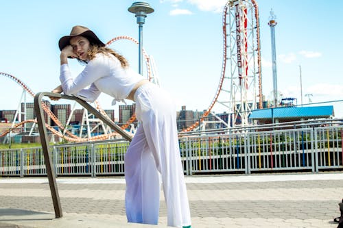 Photo of Woman in White Outfit Leaning Forward on Metal Railing with Amusement Park in the Background