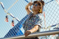 一位时尚女性戴着太阳镜，在户外的铁丝网围栏和游乐园旁摆姿势