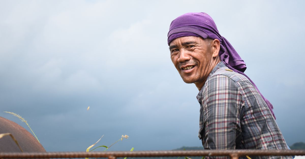 Photo by kes Belnas A smiling farmer works in a field wearing a purple bandana under a cloudy sky.