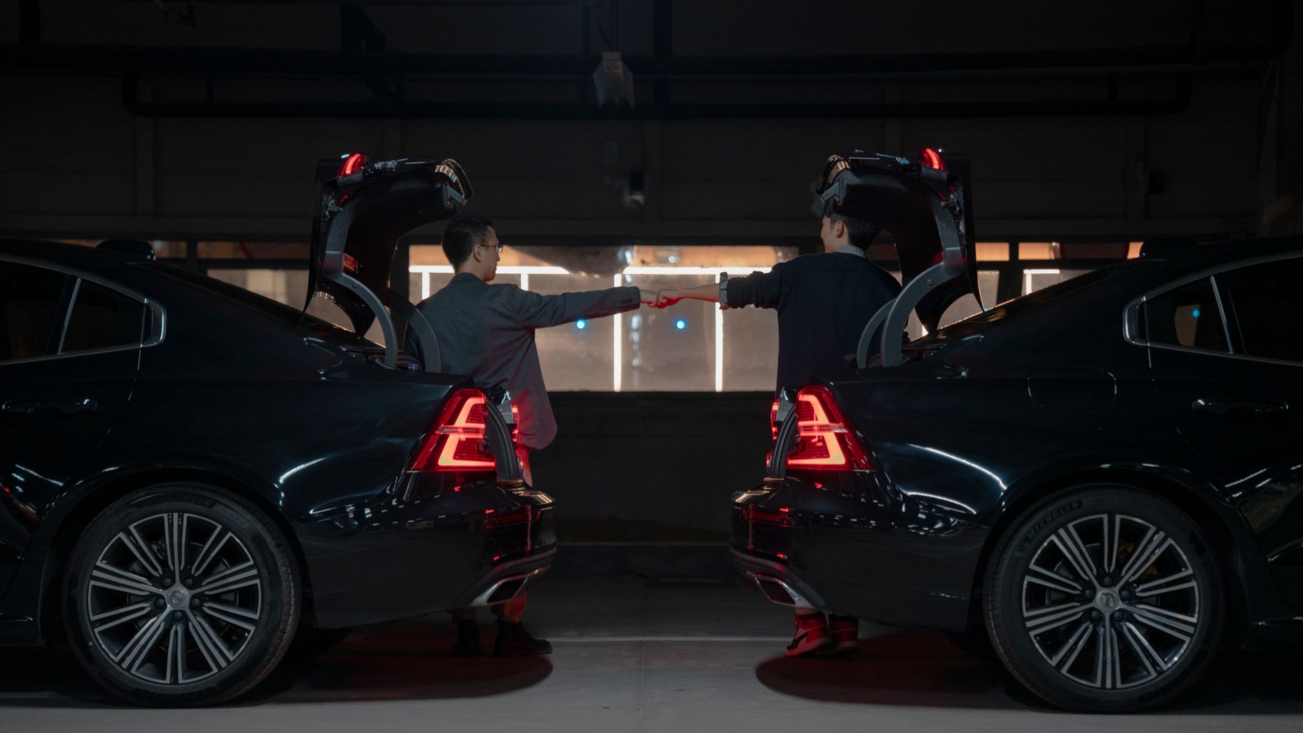 Two men in a dimly lit garage fist bumping between two cars with open trunks.
