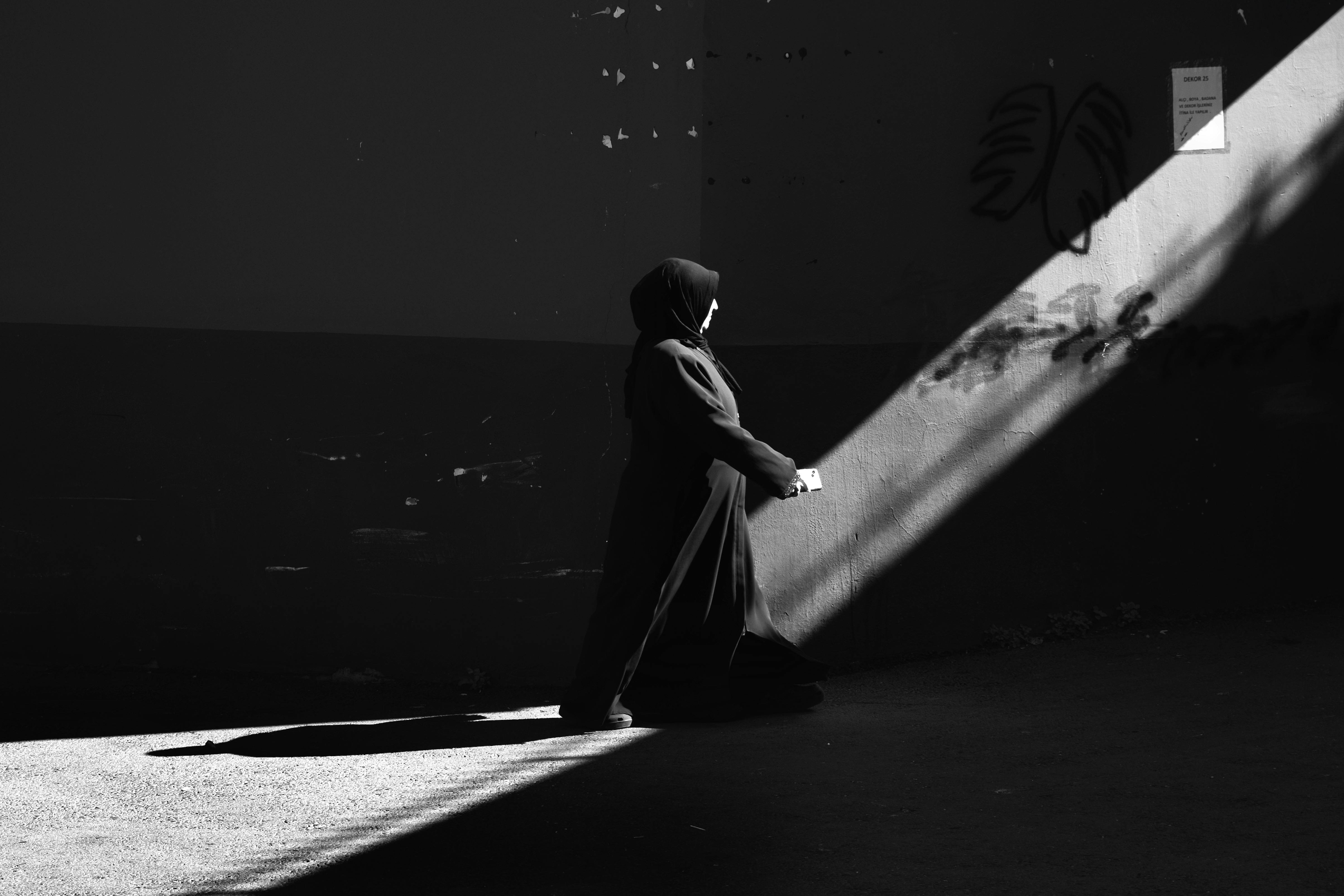Black and white image of a woman in a hijab walking through a sunlit alley.