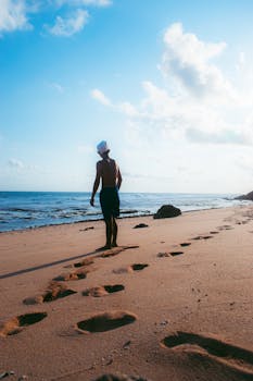 A lone person walks on a sandy beach in Yogyakarta, leaving footprints under a bright sky.