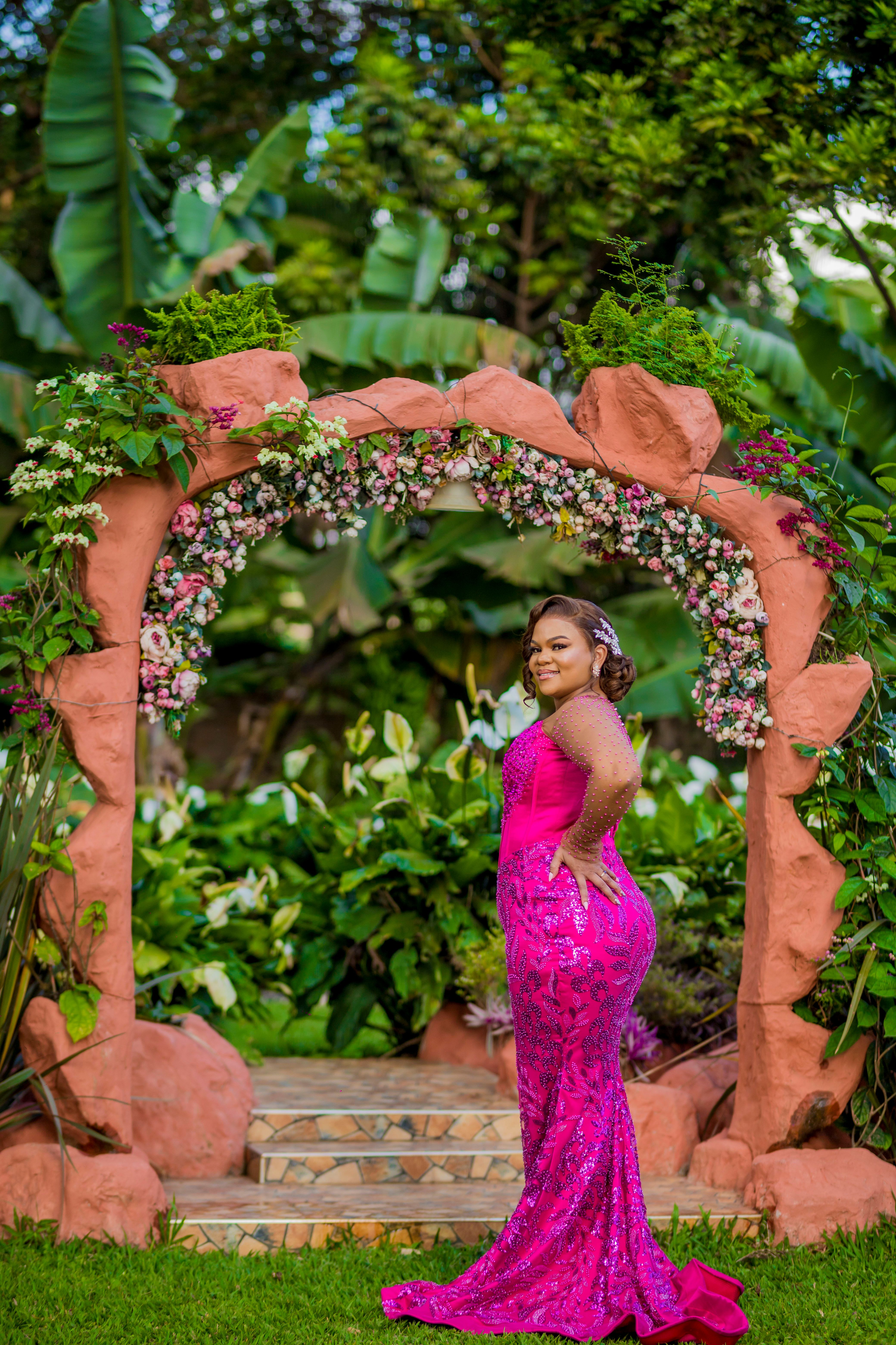 A woman in a vibrant pink dress standing gracefully under a floral arch in a lush garden setting.