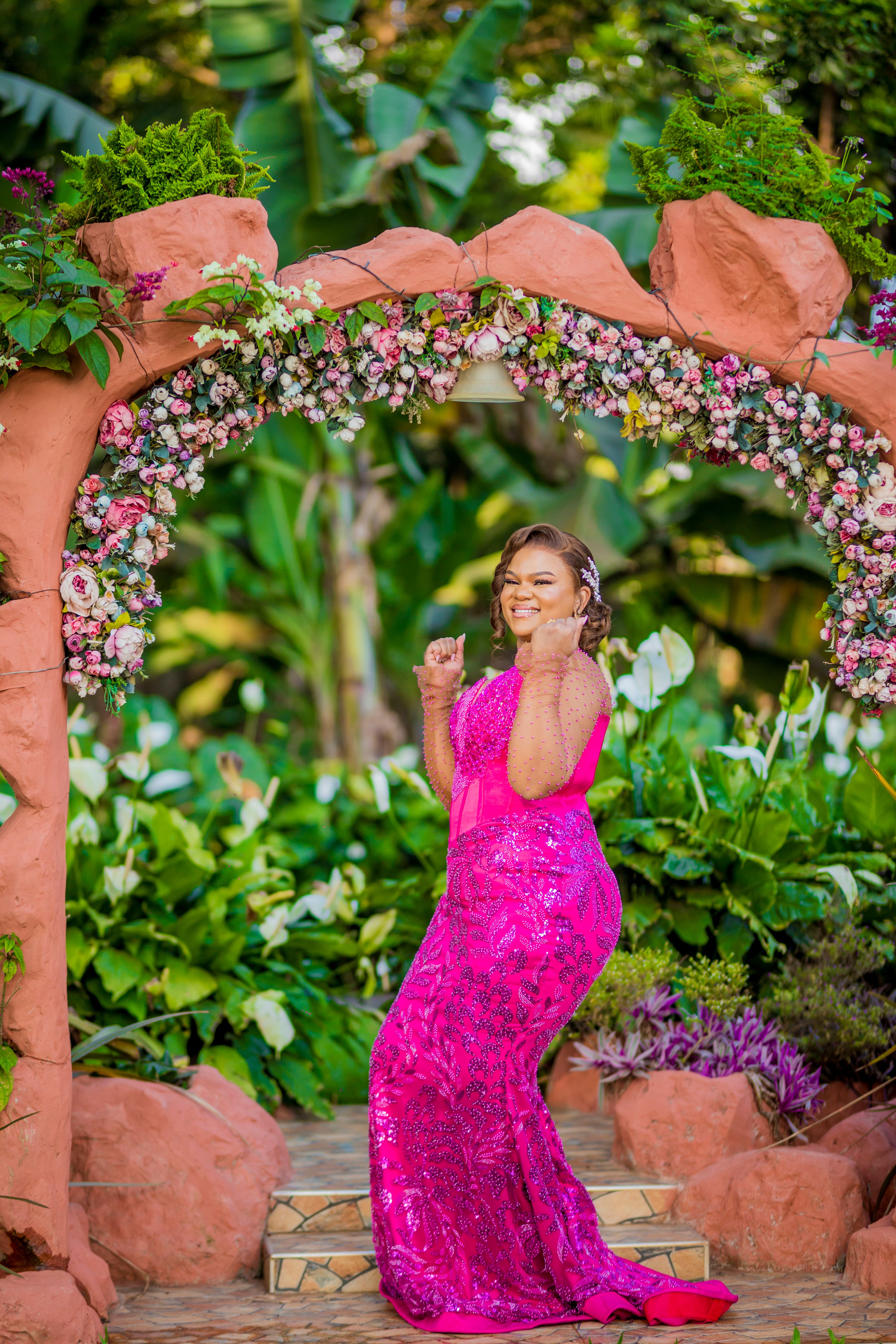 Elegant woman in a vibrant pink dress smiling under a floral arch outdoors.