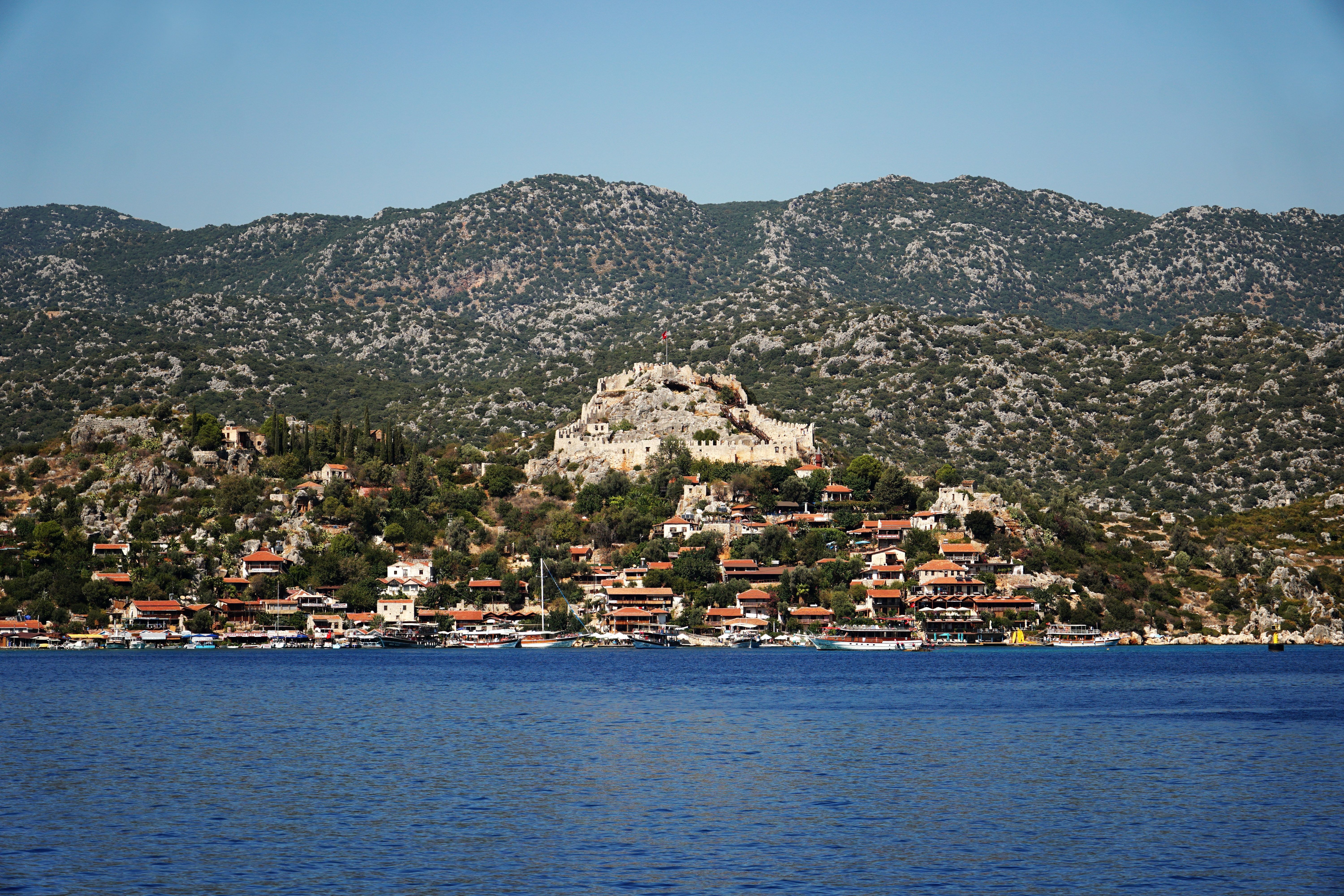A picturesque coastal village with ancient ruins against a mountainous backdrop.