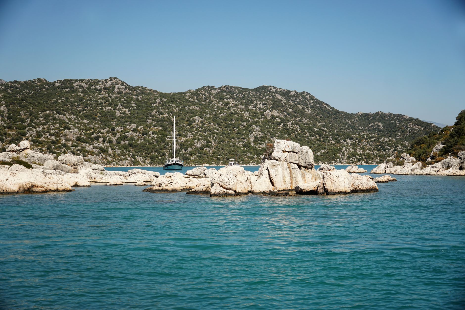 Calm sea with rocky coastlines and a sailboat against hilly terrain.